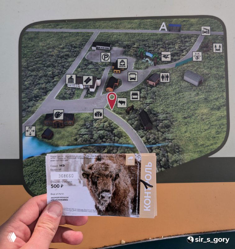 Рука держит билет и указатель с картой маршрута экскурсии «Тропа к зубрам», на фоне схемы территории питомника и значков удобств.