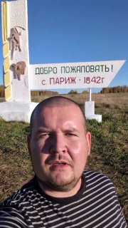Селфи автора на фоне стелы «Добро пожаловать! с. ПАРИЖ • 1842 г.» на фоне поля и деревенской дороги — панорама уральской глубинки и указатель города.