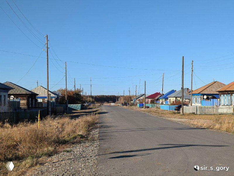 Пустая ровная улица сельского посёлка с одноэтажными деревянными домами по сторонам, показанная при дневном свете — типичная уральская глубинка.