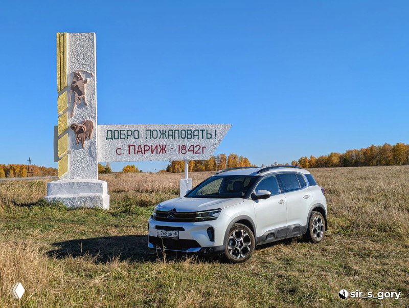 Автомобиль стоит возле монумента со стелой «Добро пожаловать! с. Париж • 1842 г.» на фоне осеннего неба — машина автора и сельский пейзаж.