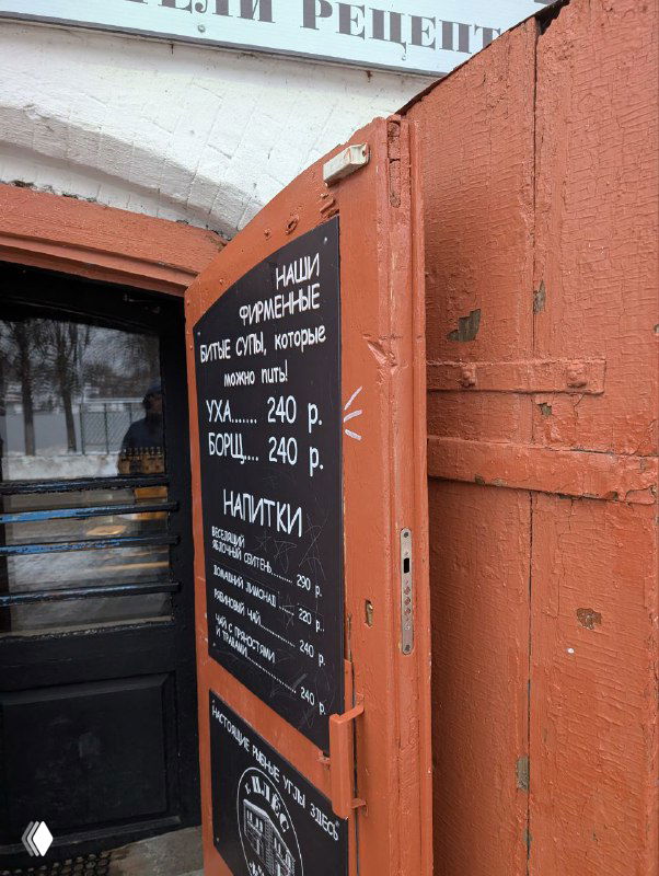 Уличная табличка с ценами на уху и напитки у входа в точку общепита, чёрная доска на фоне городского уличного пейзажа зимой.