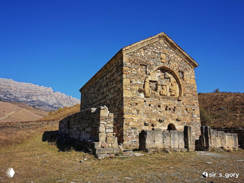 Фасад древнего каменного храма Тхаба-Ерды в Ингушетии на фоне чистого синего неба; видны арочные декоры и разрушенные участки стен.