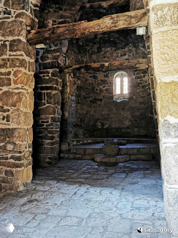 Интерьерный вид через низкую арку внутрь храма Тхаба-Ерды: каменные колонны, пол и небольшое узкое окно с мягким светом.