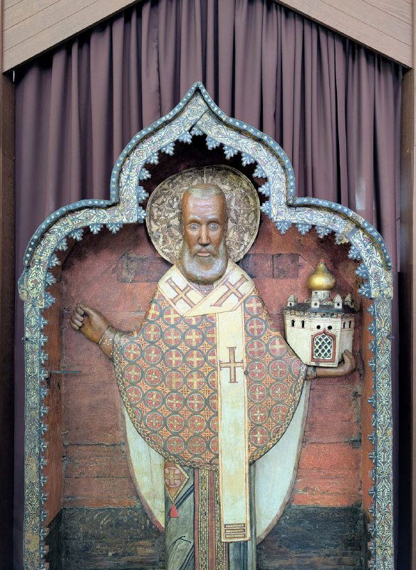 Икона в витрине музея: образ святого с орнаментированной ризой и богослужебным атрибутом, экспонируемая внутри церковного музея Каргополя.