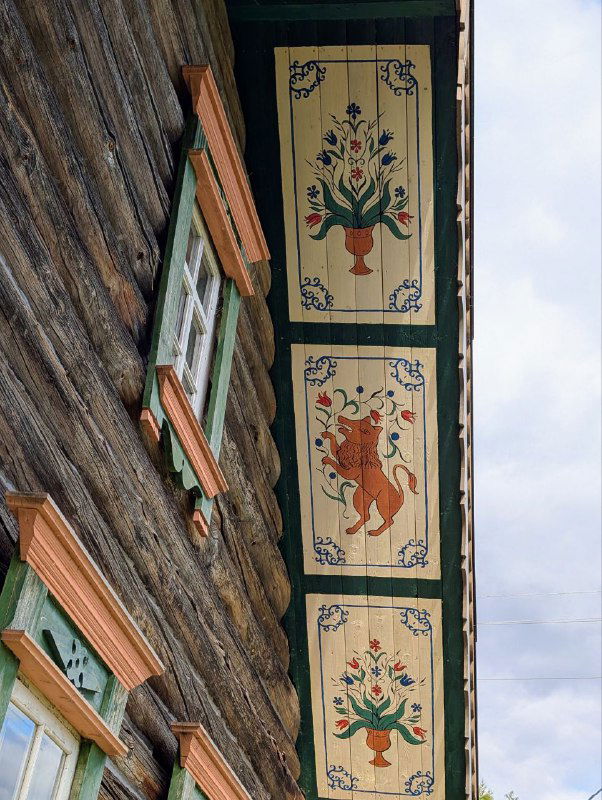 Расписной свес крыши с растительным орнаментом и фигурой зверя в традиционном стиле северной домовой росписи, вид снизу на деревянную стену и оконную раму