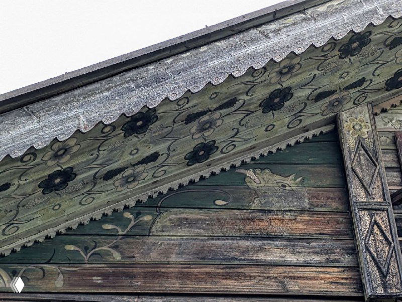 Тёмно-зелёный расписной свес с чёрными цветочными завитками и силуэтами птиц, потёртые доски и резная отделка на фасаде старого дома