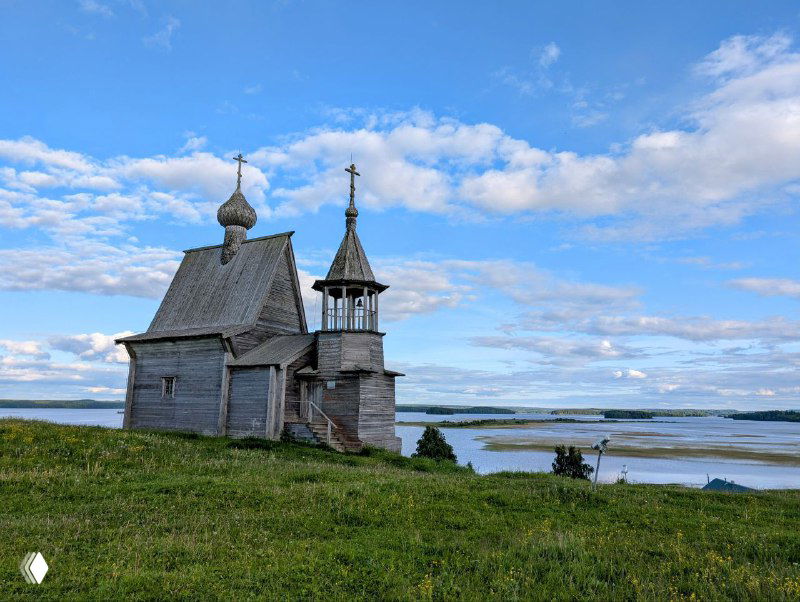 Никольская часовня Кенозерья у озера: старинная деревянная церковь на фоне зелёного склона, полей и голубовато‑облачного неба