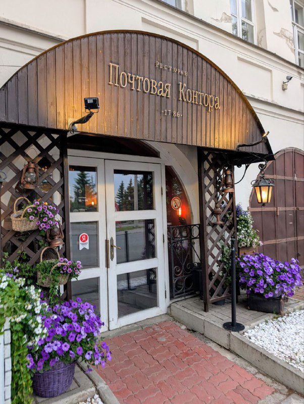 Фасад ресторана «Почтовая контора 1786» в Архангельске: деревянный козырёк над входом, цветочные корзины у двери и видимая вывеска заведения.