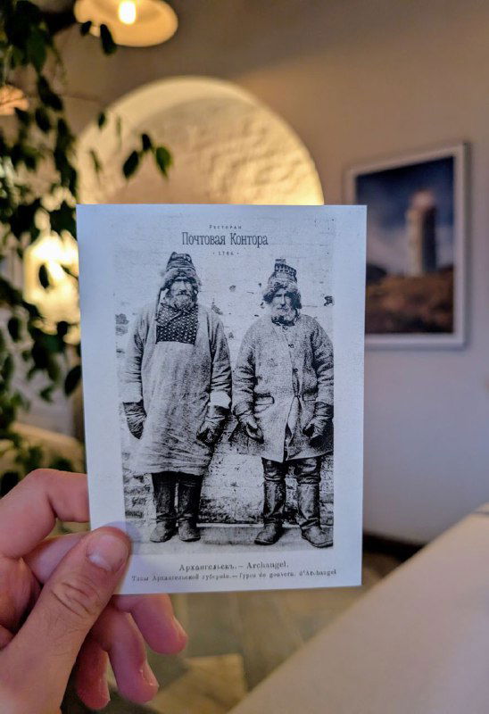 Черно-белая открытка с изображением поморов, которую держат в руке внутри зала; на заднем плане — уютный интерьер и настенные фото.