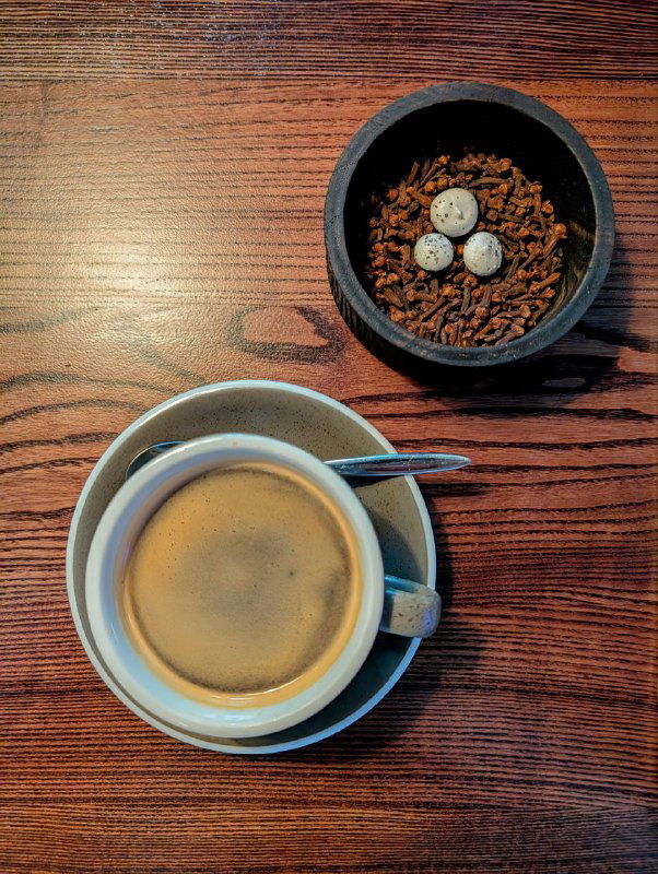 Чашка тёмного кофе в белой чашке на деревянном столе рядом с чашечкой со специями: корица, гвоздика и кардамон — кадр из ресторана в Архангельске.