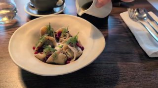 Тарелка с пельменями из устьянского кабана на тёмном столе; видно, как поливают соусом, рядом приборы и салфетка — блюдо в Roomi.