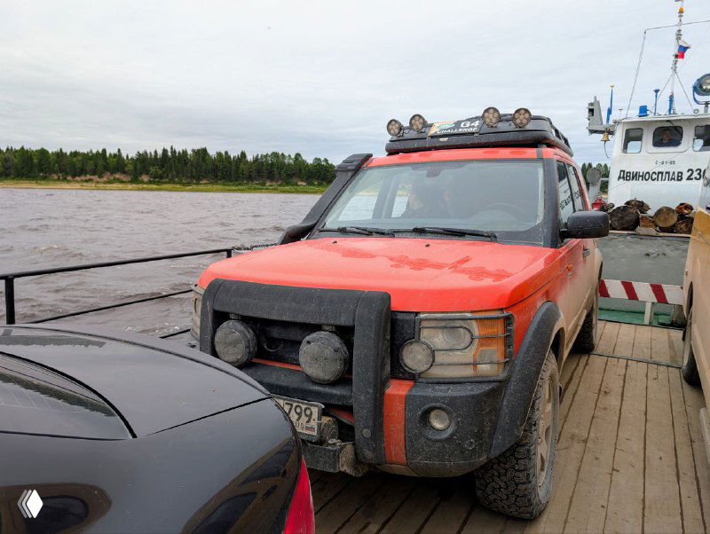 Красный внедорожник на палубе парома, ожидающего отправления по реке Северная Двина к Курострову; видны ограждения и другой причалящий корабль.