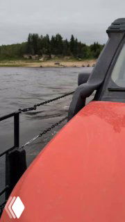 Кадр с близкого расстояния: боковая часть красного автомобиля и цепь, крепящая палубу парома, на фоне реки и дальнего берега с лесом.