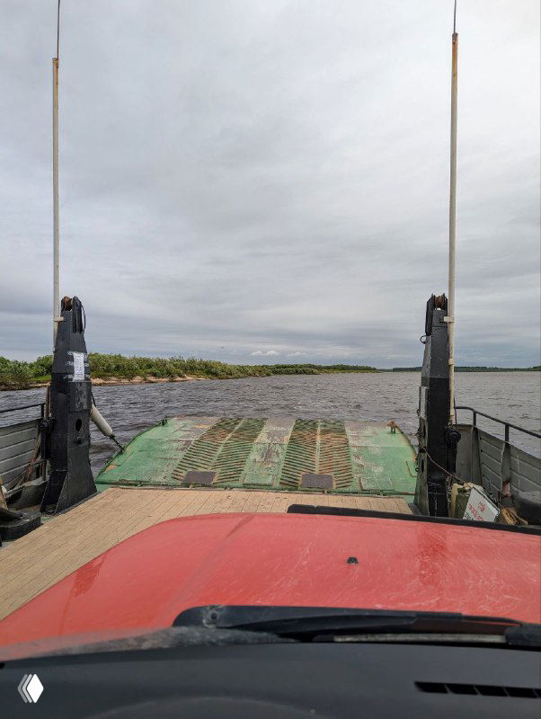 Вид с палубы парома на въездную рампу и настил, по которому подъезжает автомобиль; оттенки неба и реки под пасмурной погодой.