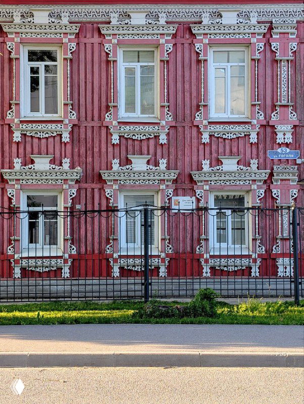 Красочный фасад дома в Вологде с белой ажурной резьбой на розово-красном фоне, ограда на переднем плане и зелёная полоса газона