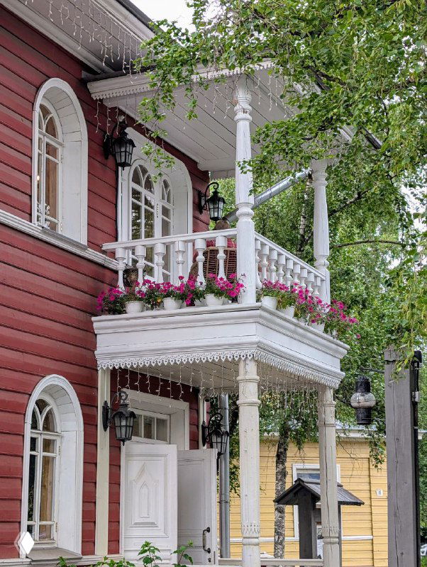 Небольший деревянный дом с белоснежным балконом-террасой над крыльцом, украшенным резными колоннами и цветочными горшками на перилах