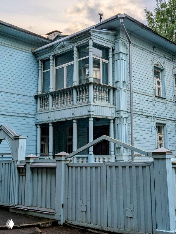 Голубой деревянный дом в Вологде с характерным закрытым балконом и резными стойками, ограждение и ворота того же тона на переднем плане