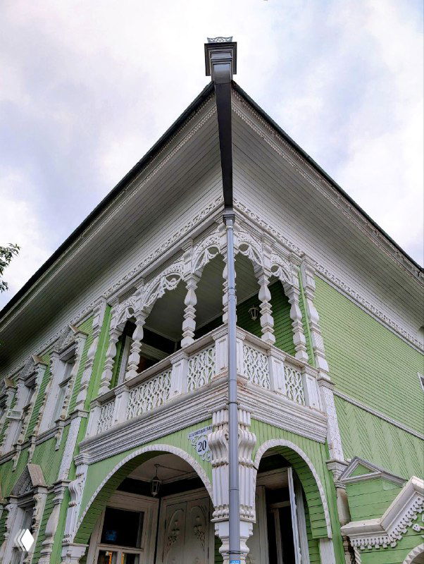 Бледно-зелёный деревянный дом с богатой ажурной резьбой на балконе и фронтоне, вид снизу подчёркивает сложный резной декор и глубину проёмов