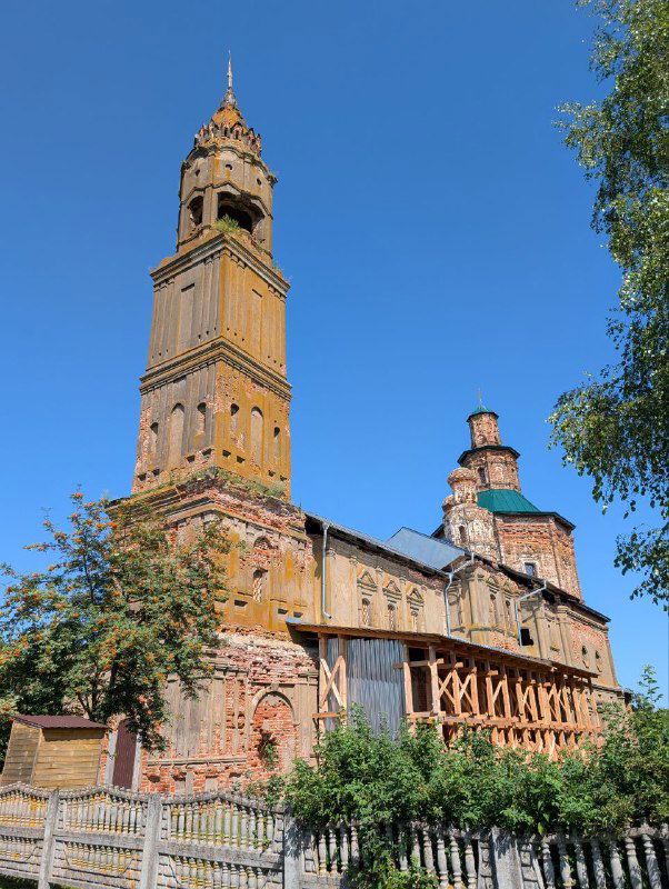 Вид крупным планом на высокую обветшалую колокольню Воскресенской церкви XVIII века на фоне ясного летнего неба и деревьев вокруг.