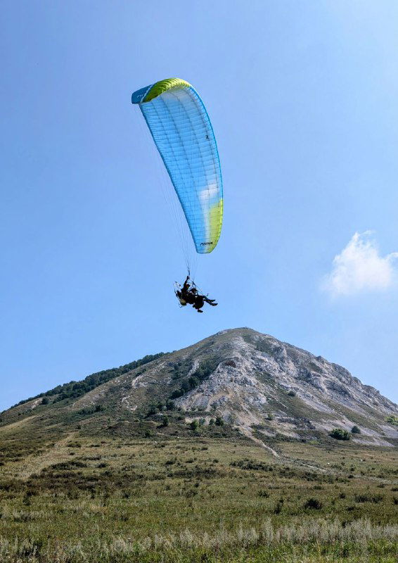 Мотопараплан в полёте над шиханом на фоне голубого неба; парапланерист и купол парашюта контрастируют с горой внизу.