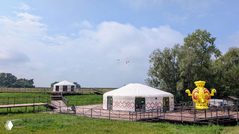 Юрты и декоративные элементы у подножия шихана: туристическая зона с деревянными ограждениями и сувенирными фигурами на зелёной лужайке.