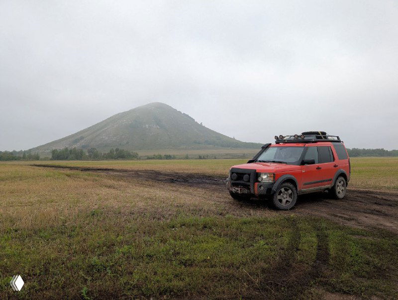 Внедорожник на грязном поле на переднем плане и шихан на заднем фоне, мокрая почва и облачное серое небо подчёркивают путешественный характер снимка.