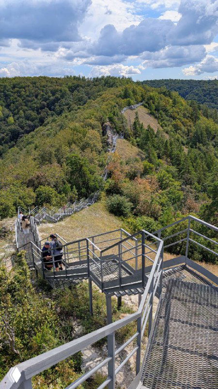 Металлическая смотровая лестница и узкий скалистый гребень (Чертов мост) с несколькими туристами на фоне лесистого хребта и реки Волга.