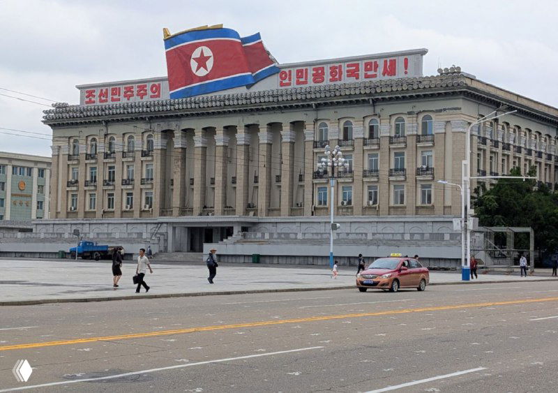 Госздание с колоннами и большим флагом КНДР на фасаде, городской проспект Пхеньяна с прохожими и автомобилем на дороге
