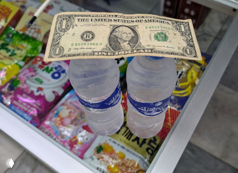 Купюра доллара на двух пластиковых бутылках воды на прилавке с разноцветными закусками — иллюстрация необходимости наличных в поездке по КНДР