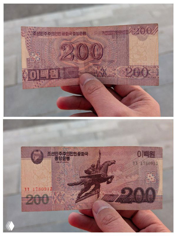 Две стороны купюры номиналом 200 вон, показанные рукой на фоне — пример корейских банкнот, которые туристам нельзя вывозить из КНДР