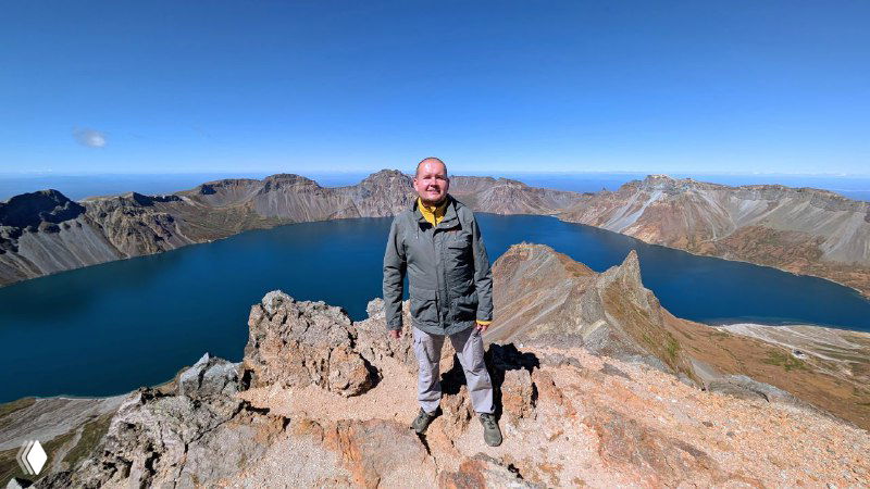 Человек стоит на краю кратера Пэкту; видно глубокое изумрудное озеро Чон, обрывистые склоны и удалённые горные пики.