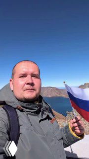 Селфи у озера Чон: турист держит флаг на фоне синей воды и скалистых хребтов кратера Пэкту под ясным небом.