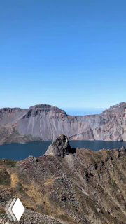 Скала и утёс на переднем плане, за ними ровная поверхность озера Чон и окружение кратера Пэкту под голубым небом.