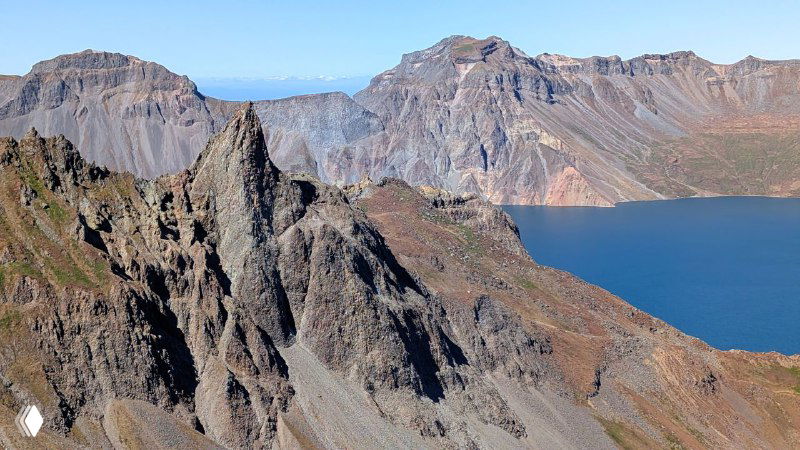 Крупный план резких базальтовых образований у края кратера Пэкту на фоне глубокого синеватого озера Чон и неба.