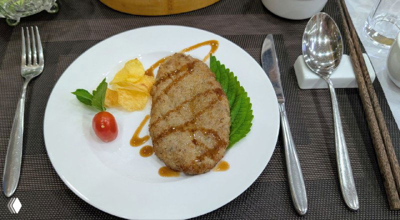 Котлета с гарниром и соусом на белой тарелке, рядом приборы и декоративный лист — часть обеда в ресторане отеля.