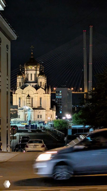 Ночная панорама Владивостока: освещённый собор на фоне висячего моста и городской улицы, вид с улицы в вечернее время.