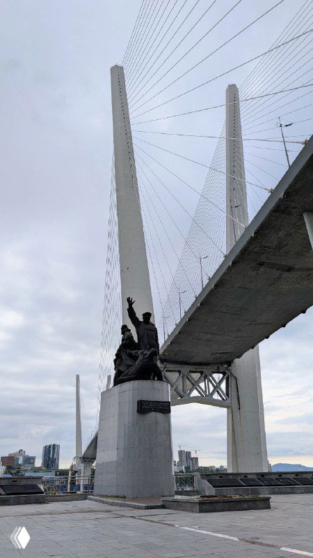 Крупный план пилонов Золотого моста и памятника Морякам торгового флота у основания, вид снизу на архитектуру и стальные тросы.