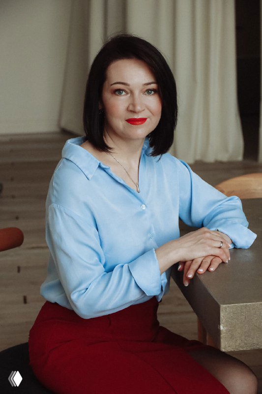 Портрет женщины в светлой блузе и красной юбке, сидящей в спокойном интерьерном пространстве офиса; деловой профильный портрет для страницы автора канала.