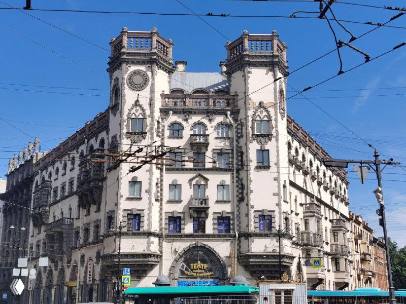 Фасад крупного петербургского доходного дома начала XX века с богатым архитектурным декором, окнами и трамвайными проводами на фоне чистого неба.