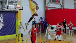 Фрагмент матча: борьба под корзиной между командами в белой и красной форме, судьи и баннеры турнира видны на заднем плане.