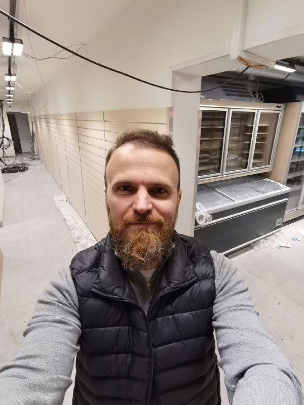 Селфи автора на фоне торгового зала и холодильных блоков: мужчина в жилетке, видна внутренняя отделка торгового помещения и оборудование магазина.