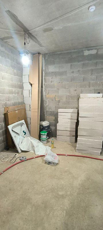 Пустое помещение ремонтного этапа: стопки газоблока, ведро, упаковки смесей и оголённые провода под потолочной лампой.