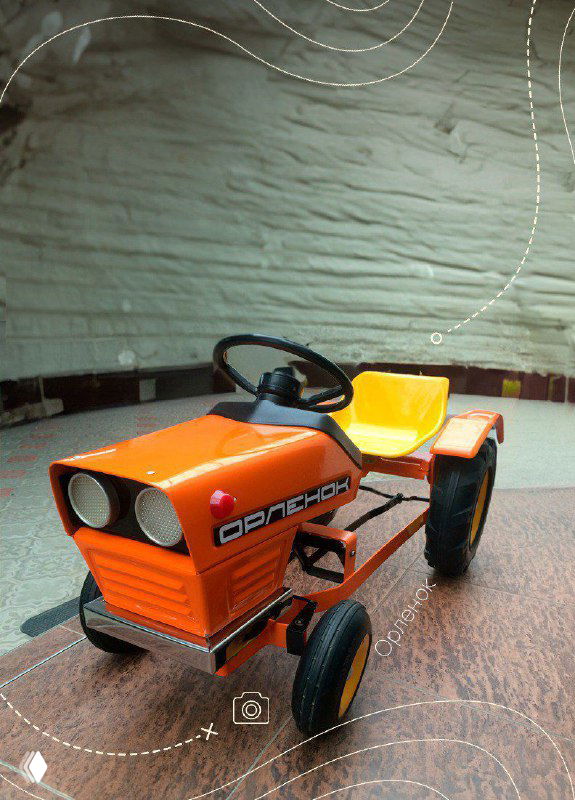 Оранжевый педальный трактор с металлической рамой и большими колёсами, снимок на деревянном полу подчёркивает индустриальный характер игрушки.