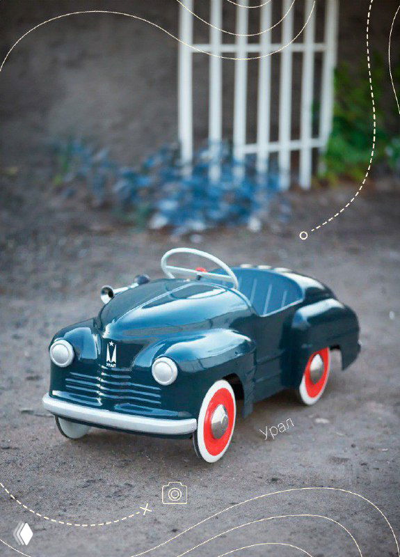 Тёмно‑синяя педальная ретро‑машина с округлым кузовом и белыми колёсами, фото в уличной обстановке демонстрирует классический дизайн игрушки.