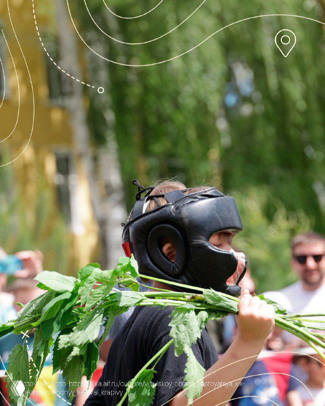 Участник в защитном шлеме держит ветки крапивы на фоне зрителей и деревьев — кадр крапивного боя на народном празднике в селе Крапивна.