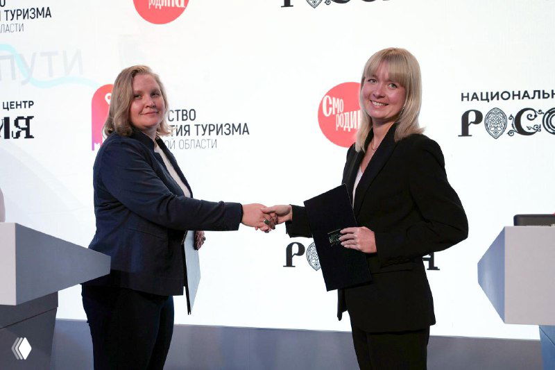 Две женщины пожимают руки на сцене во время подписания соглашения: видна церемония, кулиса с логотипами выставки «Регион‑2030» и надпись Национального центра «Россия».