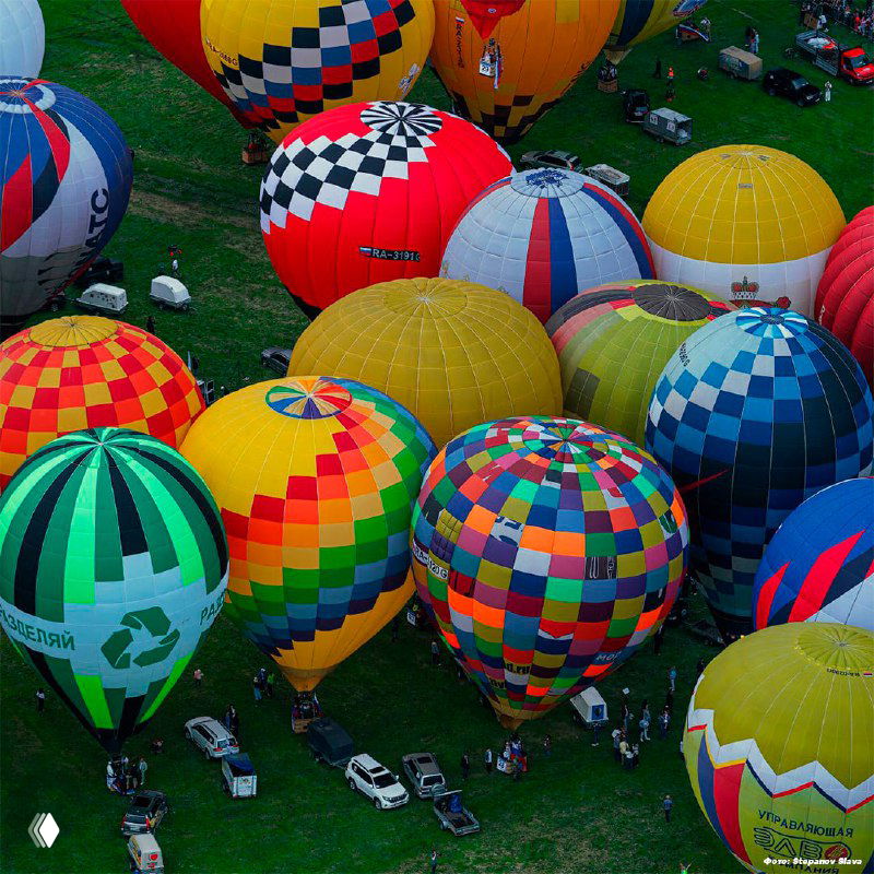 Яркие разноцветные воздушные шары, собранные на поле фестиваля в Великих Луках; сверху видны люди, машины и подготовка к взлету.