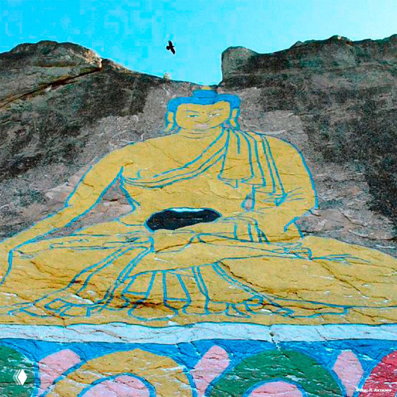 Крупный план цветного изображения Будды Шакьямуни, нанесённого на скалистую стену горы Баян-Хонгор; вид снизу, вокруг заметны сосны и камень.