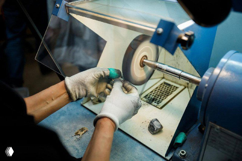 Процесс обточки и полировки украшения на шлифовальном станке: руки в перчатках удерживают деталь под светом в цехе ювелирного завода.
