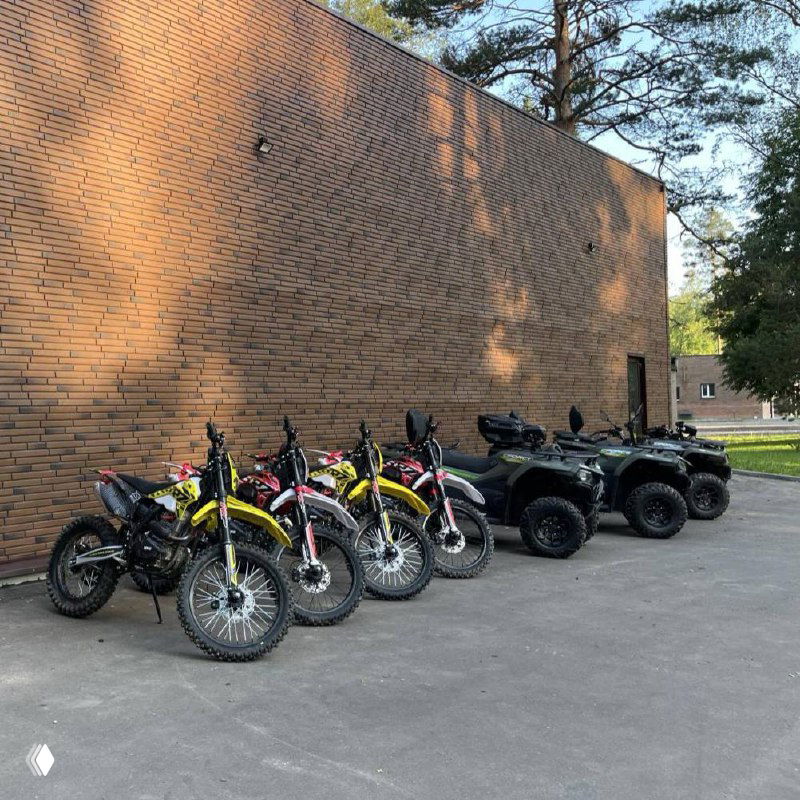 Несколько внедорожных мотоциклов и квадроциклов припаркованы у кирпичного здания перед началом маршрута и подготовки к заезду.